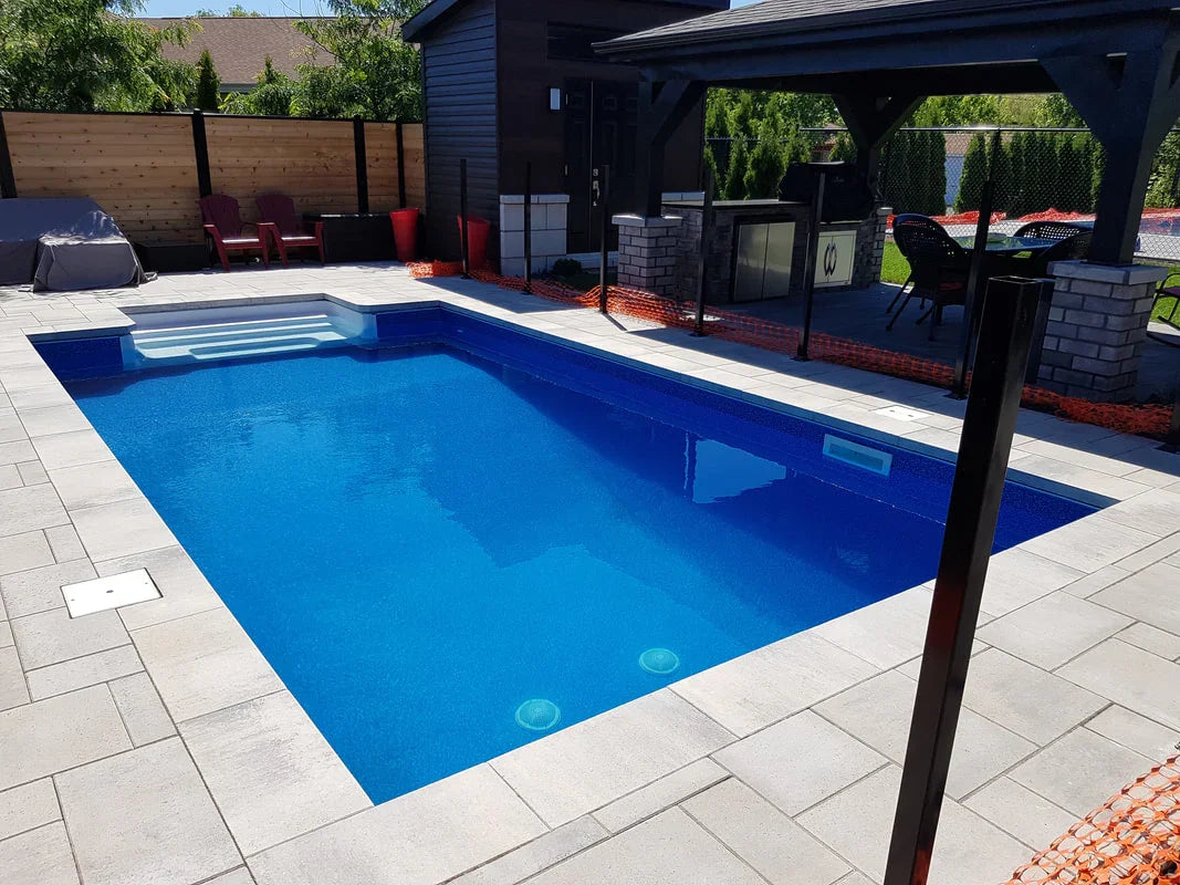 Piscine creusée moderne avec terrasse en pavé, coin détente et pool house en arrière-plan.
