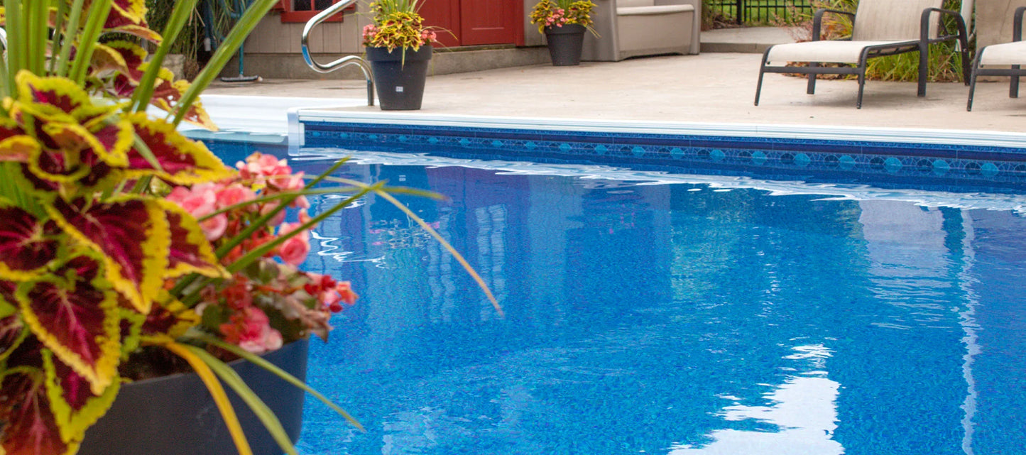 Piscine creusée bleue avec margelle, fleurs en pot et chaises longues sur terrasse extérieure.