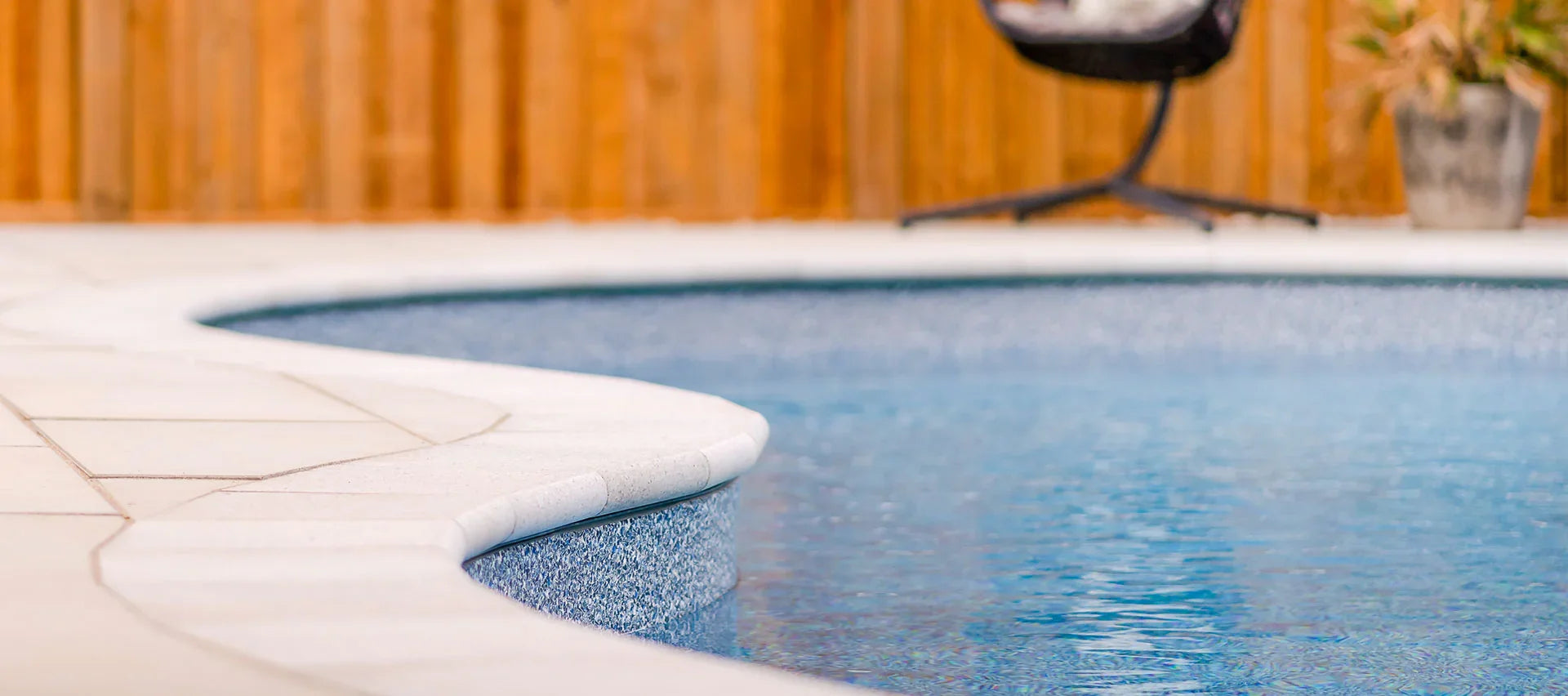 Bord de piscine résidentielle avec margelles claires et eau bleue, clôture bois en arrière-plan
