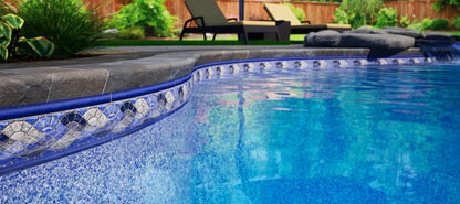 Rebord de piscine creusée avec margelle en pierre, eau bleue et décor de jardin