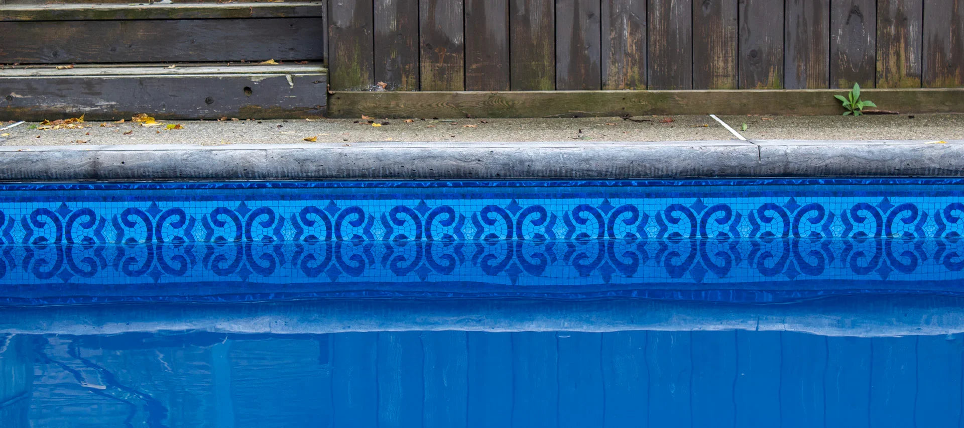 Bord de piscine bleu avec liner à motif géométrique, margelle en béton et clôture en bois