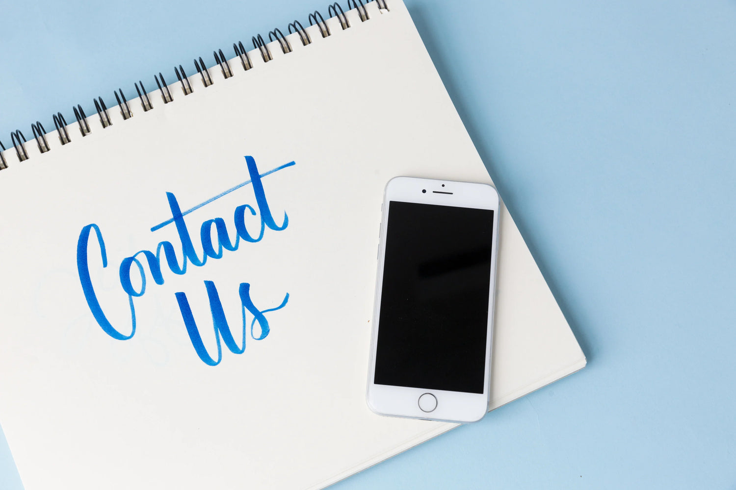 Smartphone et carnet avec texte « Contact Us » sur fond bleu, support contact piscine.