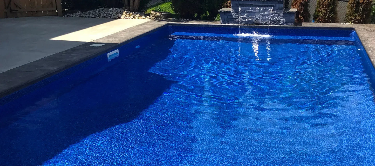 Piscine creusée extérieure avec margelles grises et fontaine murale, eau bleue scintillante