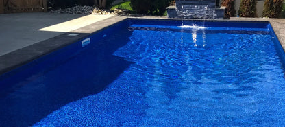 Piscine creusée extérieure avec margelles grises et fontaine murale, eau bleue scintillante
