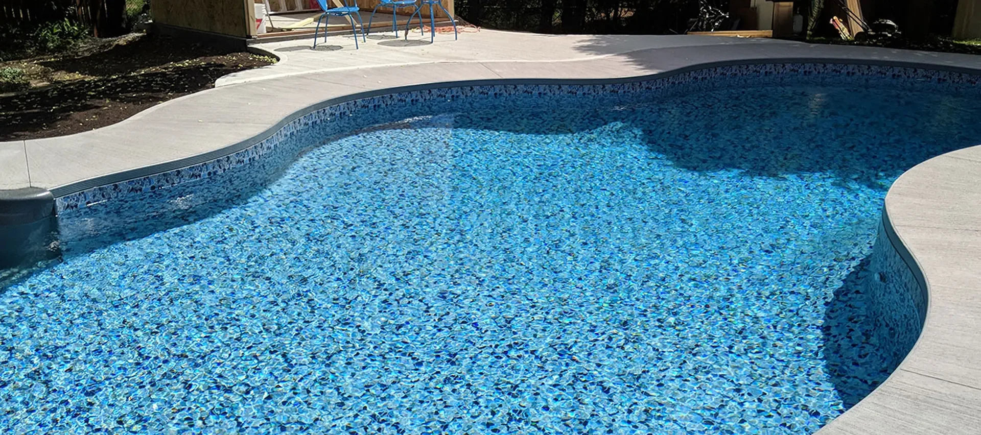 Piscine extérieure moderne avec finition en mosaïque bleue et terrasse en béton, ensoleillée.