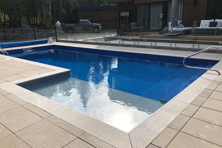 Piscine creusée moderne avec margelles en pierre, terrasse en dalles et rambarde en verre.